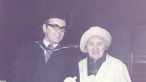 Barry in his graduation gown and hat with his grandmother. They are smiling together.